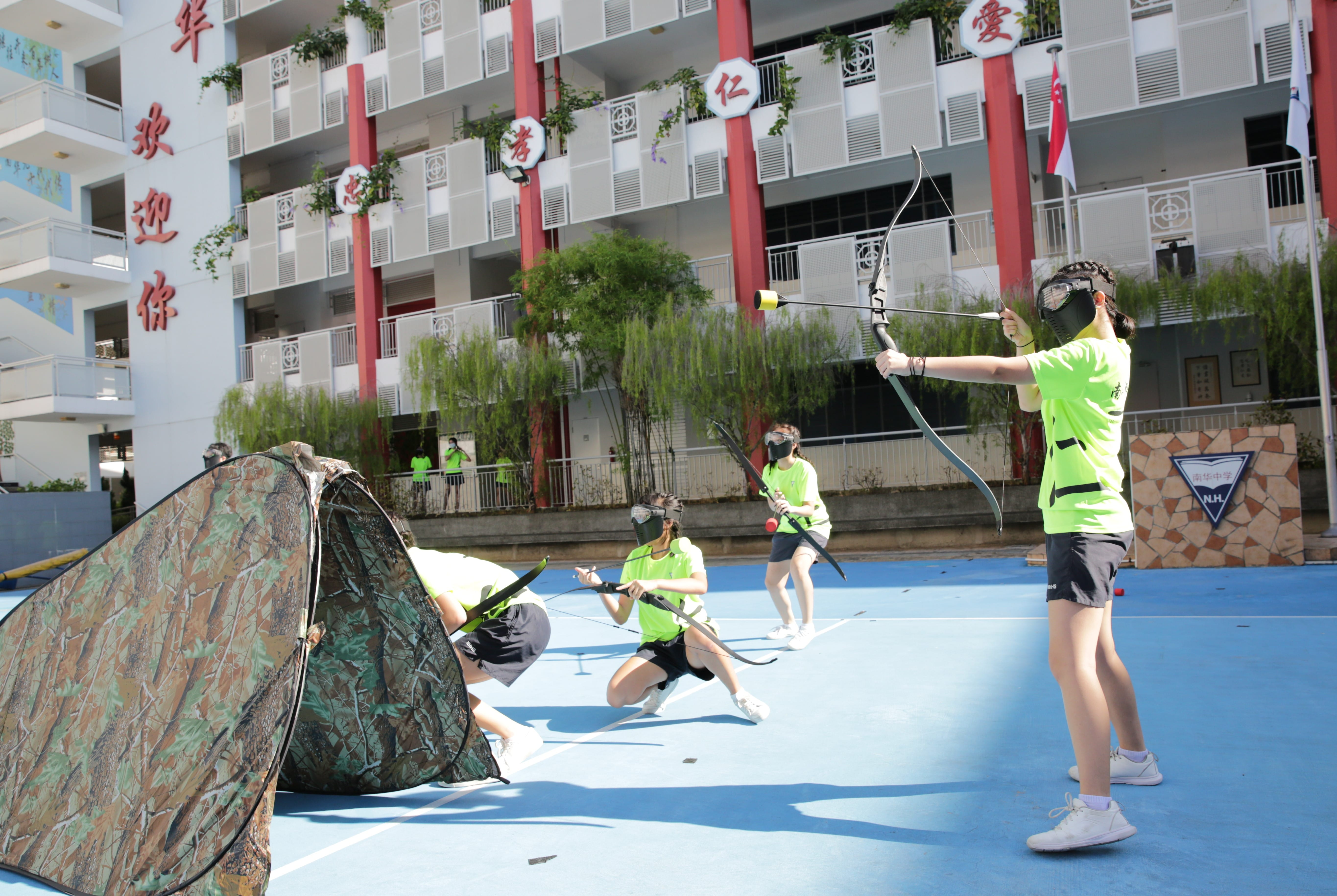 Students in green shirts and face masks play archery tag on a blue court near a camouflage tent, with a school building behind.